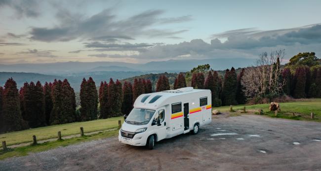 White campervan with red and yellow stripes parked overlooking a valley with distant mountains.