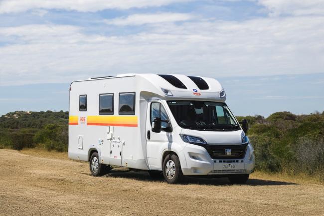 White Fiat campervan with yellow and orange side stripes, parked on a dirt track under a partly cloudy sky.