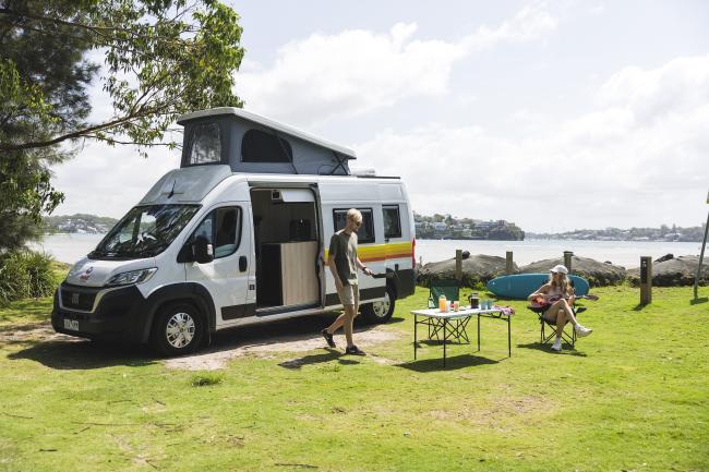 Couple camping on grass beside water next to a white pop-top campervan with its side door open.