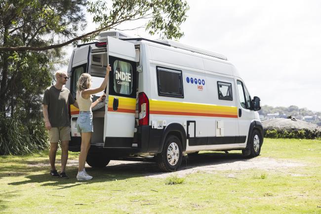 A couple stands by the open back doors of a white Indie Campers campervan parked on a grassy field.