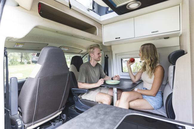 A man and woman sit at a dinette table inside a campervan interior, smiling and eating.