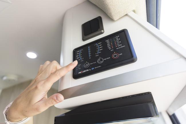 A hand operating a digital control panel displaying battery and water levels inside a campervan.