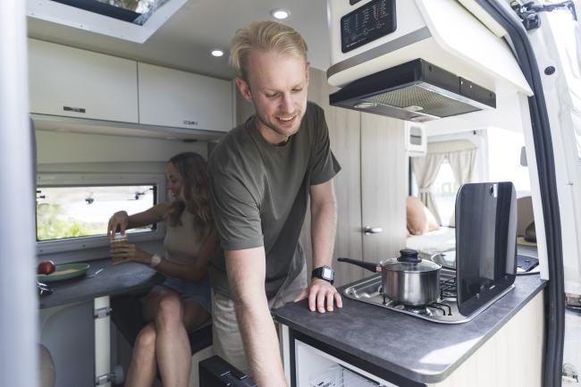 Man reaching into a fridge inside a campervan kitchen, with a woman sitting at a table in the background.