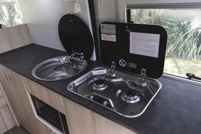 Compact campervan kitchen with a stainless steel sink and two-burner gas stove, both with open black glass lids.