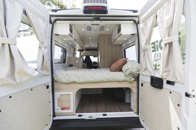 Campervan interior through open rear doors, featuring a raised bed with green patterned bedding and under-bed storage.