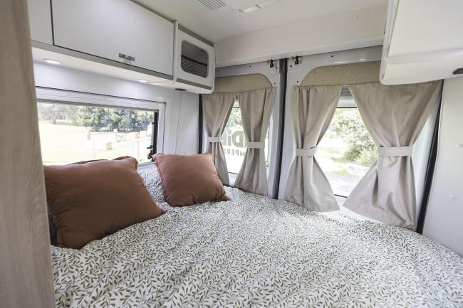 Double bed inside a campervan with a leaf-patterned duvet, brown pillows, and rear window curtains.