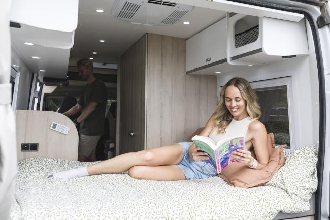 Woman reading a book on a bed inside a campervan with a man standing in the background kitchen area.
