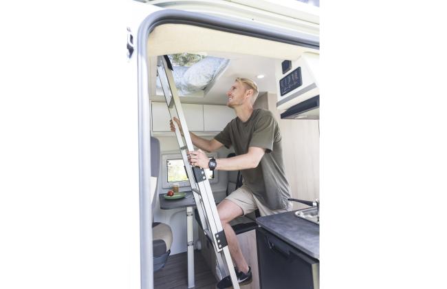 A man climbs a metal ladder inside a campervan towards the overhead bed area.