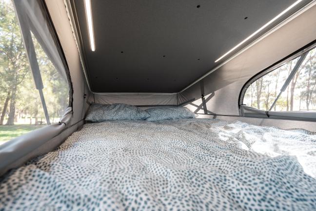 Double bed with patterned bedding inside a campervan pop-top roof tent, lit by LED strips with views of trees.