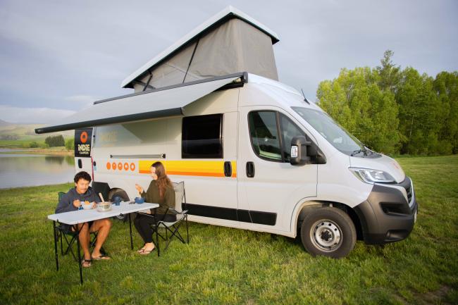 Couple dining outside a white campervan with an open pop-top roof and extended awning on a grassy field by a lake.