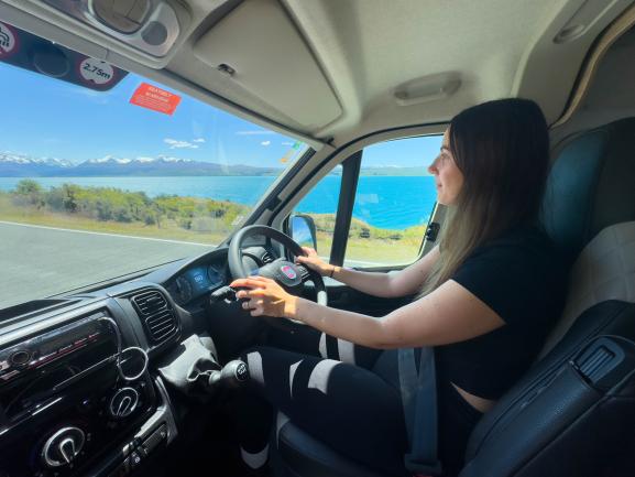 Woman driving a campervan on a road alongside a vibrant blue lake with snow-capped mountains in the background.