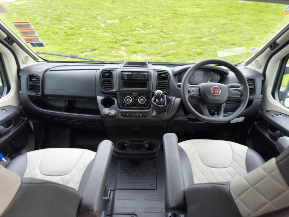 Right-hand drive Fiat campervan cab featuring two-tone grey and cream quilted seats, a black dashboard and steering wheel.