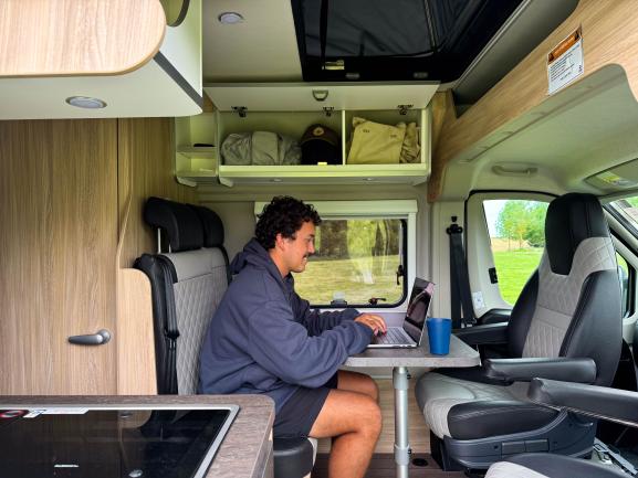 A man sits at a table inside a campervan, working on a laptop next to a blue cup.