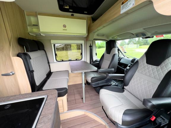 Campervan interior with grey quilted swivel seats, a central dining table, rear bench, and light timber-look finishes.