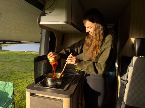 Woman cooking inside a campervan, pouring tomato sauce into a pot on a stove with a green field visible outside.