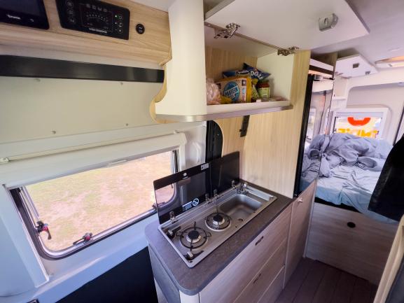 Campervan interior featuring a compact kitchen with gas burners and sink, open overhead cabinet, and bed in the background.