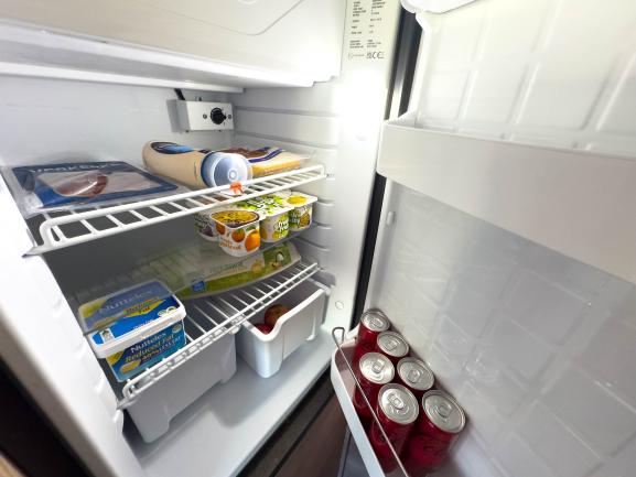 Interior of a compact campervan fridge stocked with canned drinks, spreads, yoghurt, eggs, and fresh produce.