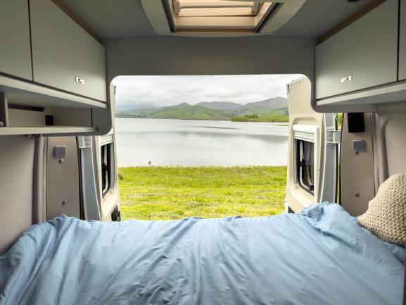 View from inside a campervan bed looking through open rear doors to a lake and green hills.