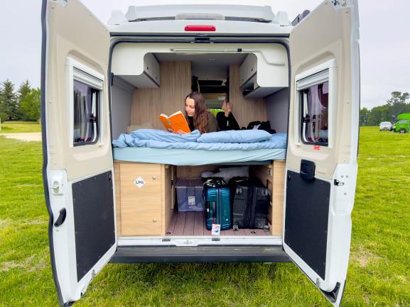 Woman reading on a bed inside a campervan with open rear doors, showing suitcases stored in the compartment below.
