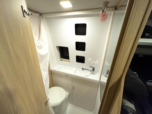 Compact campervan bathroom with a cassette toilet, small basin, tap, wall shelving recesses, and timber-look cabinets.