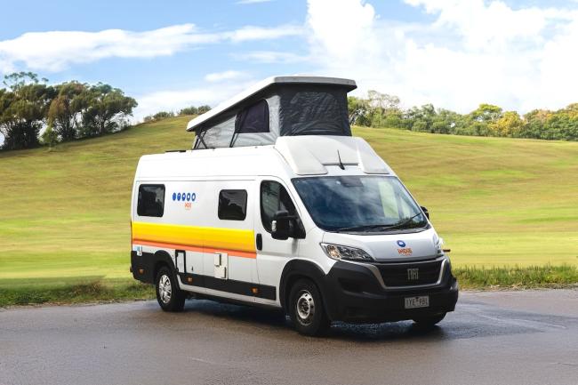 White campervan with a raised pop-top roof tent parked on a paved road against a grassy hill.