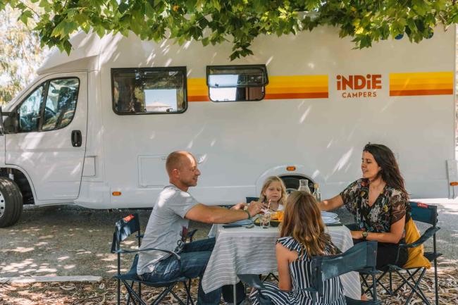 Family dining at an outdoor table beside a white Indie Campers campervan in the shade.