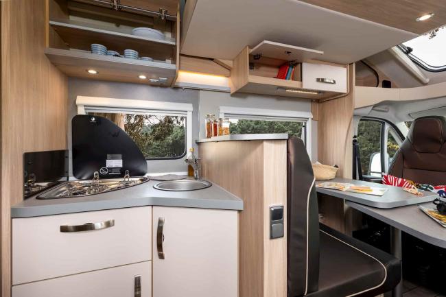 Interior of a campervan showing a compact kitchen with sink and stovetop, and an adjacent dining area with booth seating.