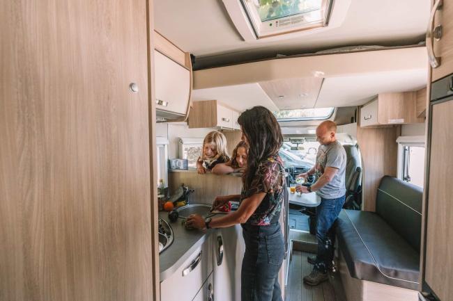 Family of four inside a campervan as a woman prepares food at the kitchen bench and a man pours a drink.
