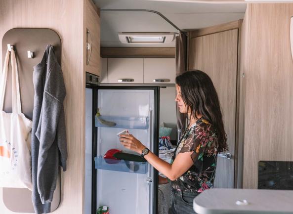 A woman inside a campervan looks at a food container from an open refrigerator.
