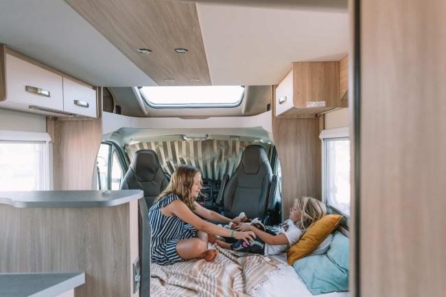 Two girls play on a bed inside a campervan interior with light-coloured cabinets and cab seats in the background.