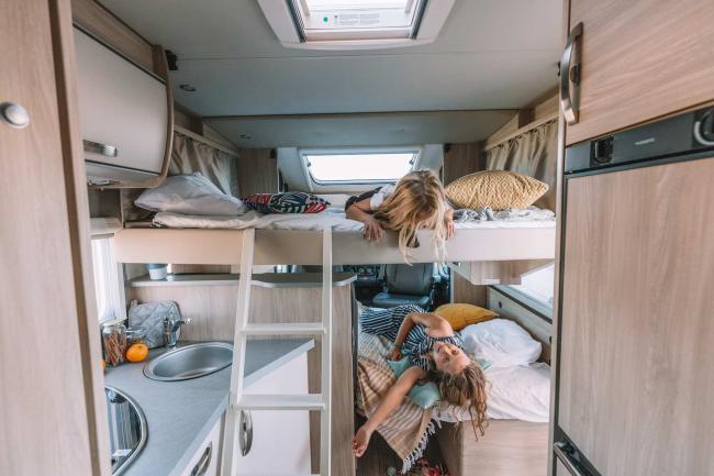 Two children on bunk beds inside a campervan with a small kitchen and ladder.