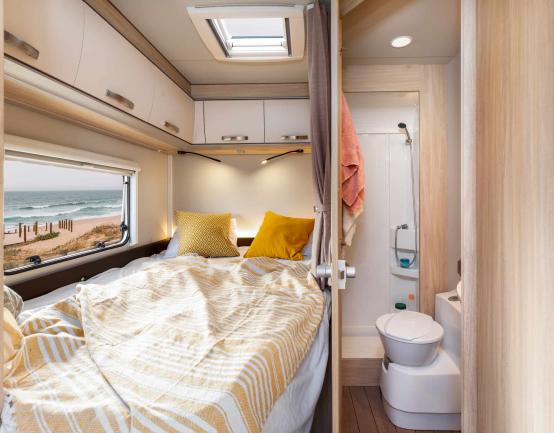 Campervan interior showing a bed beside a window with a beach view, next to a compact bathroom with a toilet and shower.
