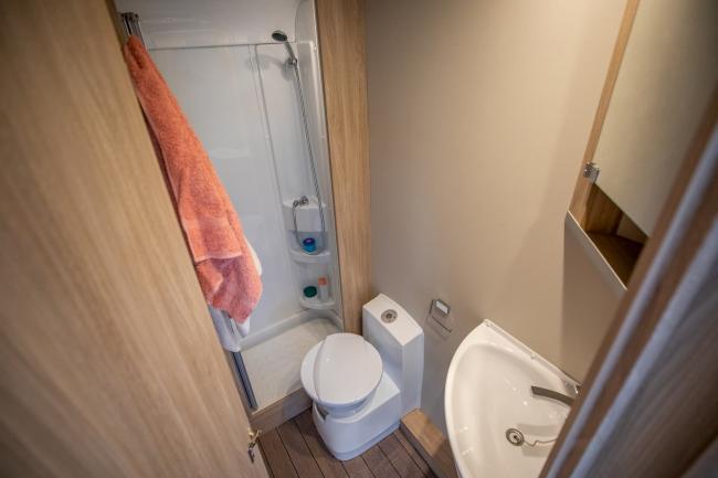 Compact campervan bathroom featuring a small toilet, corner sink, timber panelling, and a shower with a hanging towel.