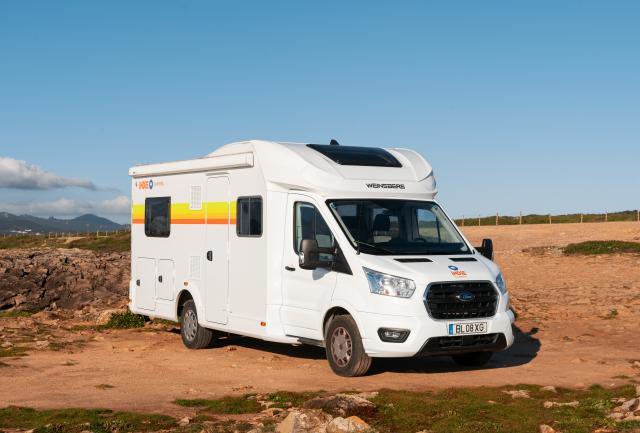 White Weinsberg campervan with yellow and orange stripes parked on dirt ground under a clear blue sky.