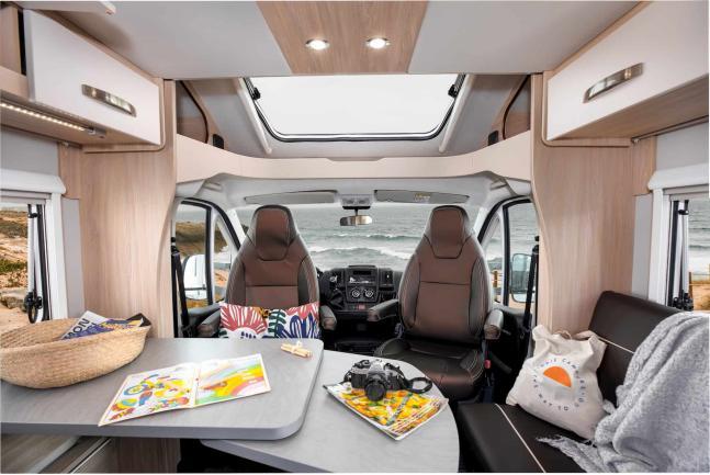 Campervan interior showing swivel seats facing a dining table, with an ocean view visible through the front windscreen.