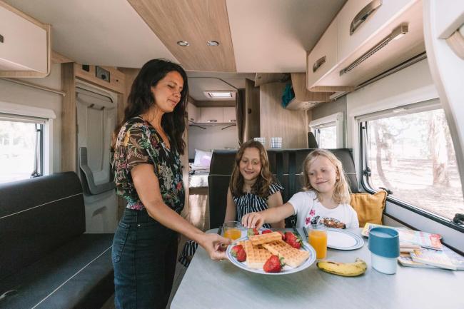 A woman serves waffles to two young girls sitting at a dining table inside a campervan.