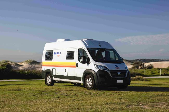 White Fiat campervan with yellow and orange stripes parked on a grassy field.
