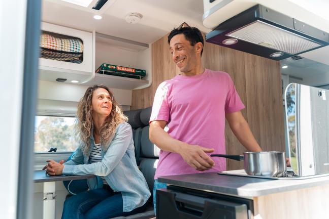 A man in a pink t-shirt stands by the stove and smiles at a woman sitting at the dining table inside a campervan.