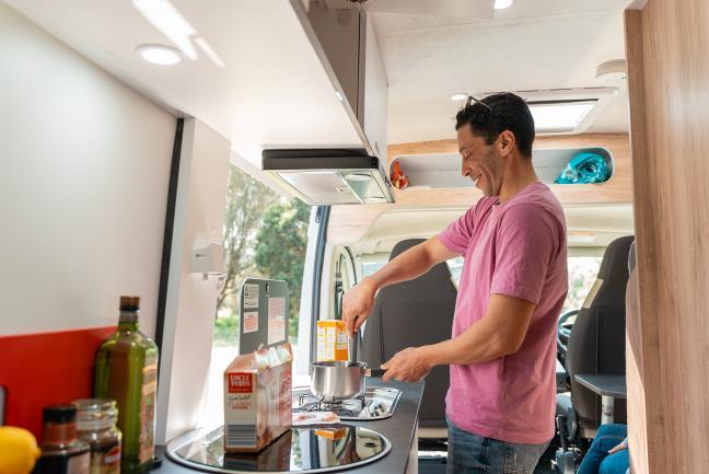 Man stirring a pot on a small stove inside a campervan kitchen.