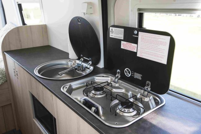 Compact campervan kitchen with a round stainless steel sink and a two-burner gas stove, both with open black lids.