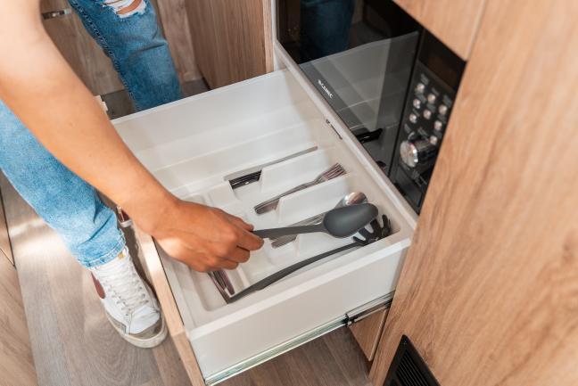 A person reaches into an open cutlery drawer with a white organiser, built into timber-look campervan cabinetry.