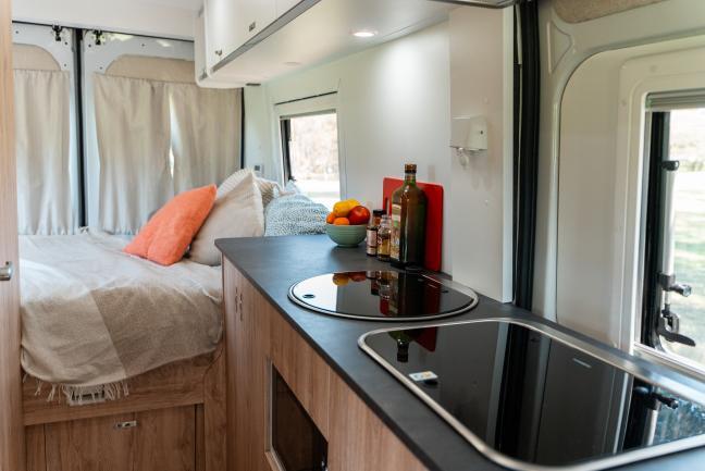 Campervan interior showing a timber kitchen benchtop with appliances next to a bed with pillows.