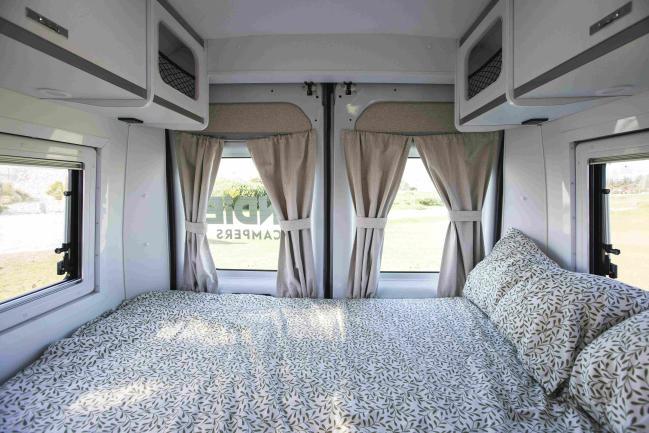 Campervan interior showing a bed with leaf-patterned linen, overhead cabinets, and rear windows with beige curtains.