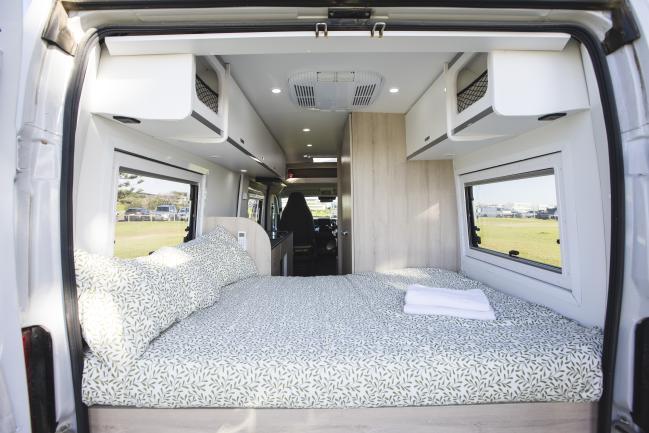 Interior of a campervan featuring a double bed with leaf-patterned sheets, white overhead cabinets, and side windows.