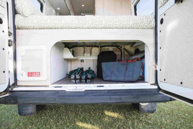 Open rear doors of a campervan showing under-bed storage filled with bags, camping chairs, and a gas storage hatch.
