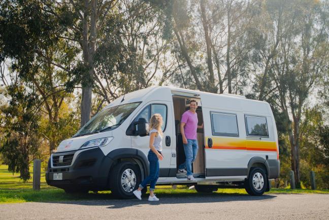 A couple stands next to a white campervan with yellow and orange stripes, parked near tall trees.