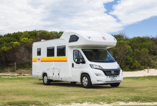 White Fiat campervan with yellow and orange stripes parked on a grassy field.