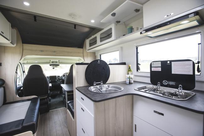 Compact campervan kitchen with a sink, gas stove, light timber-look cabinets, and front cabin seats visible.