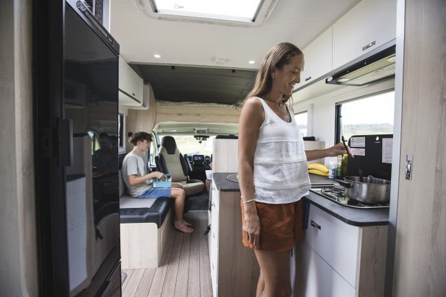 Inside a campervan, a woman stirs a pot on the kitchen stove while a teenager sits on a bench seat in the background.