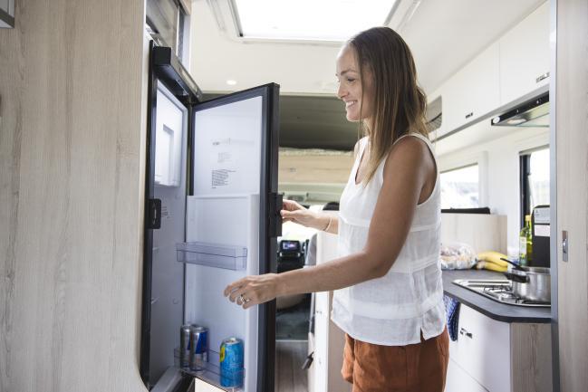 Smiling woman looks inside a compact refrigerator in a campervan kitchen.
